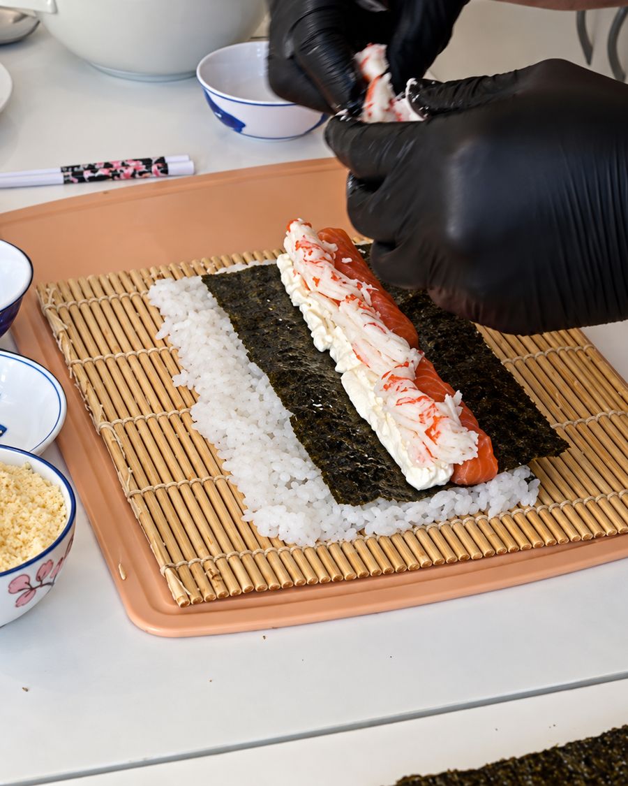 Manos con guantes armando un roll sobre la esterilla de bambú, con el arroz y el nori extendidos.